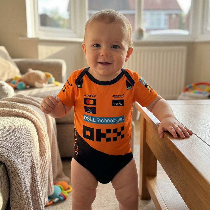 Smiling baby standing in McLaren F1 Longsleeve Jumpsuit 2025 with OKX and sponsor logos in bright home living room