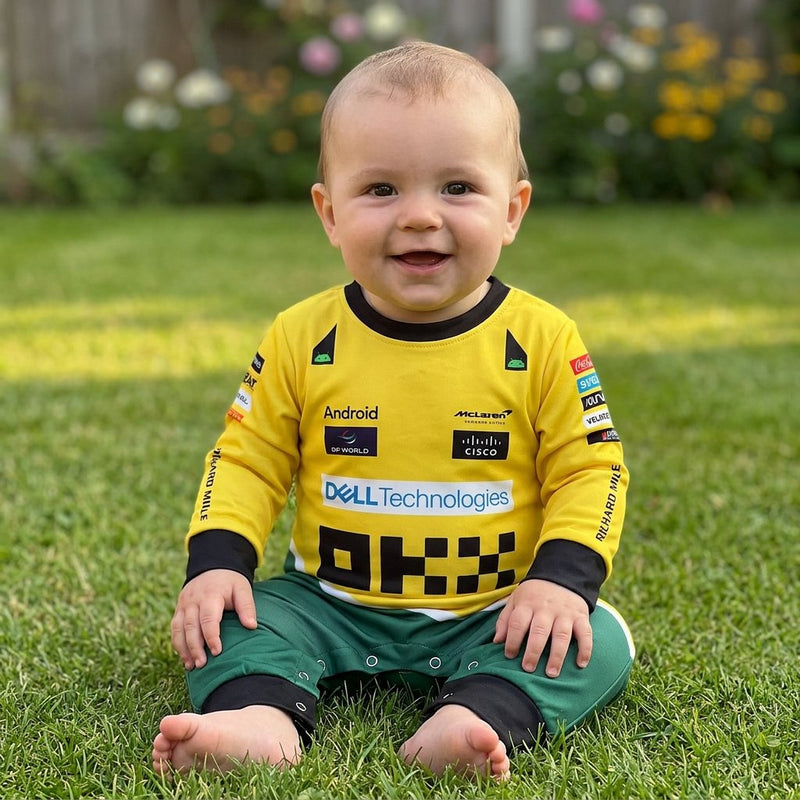 Smiling baby on garden grass wearing a yellow and green McLaren Senna F1 replica racing suit onesie with sponsor logos