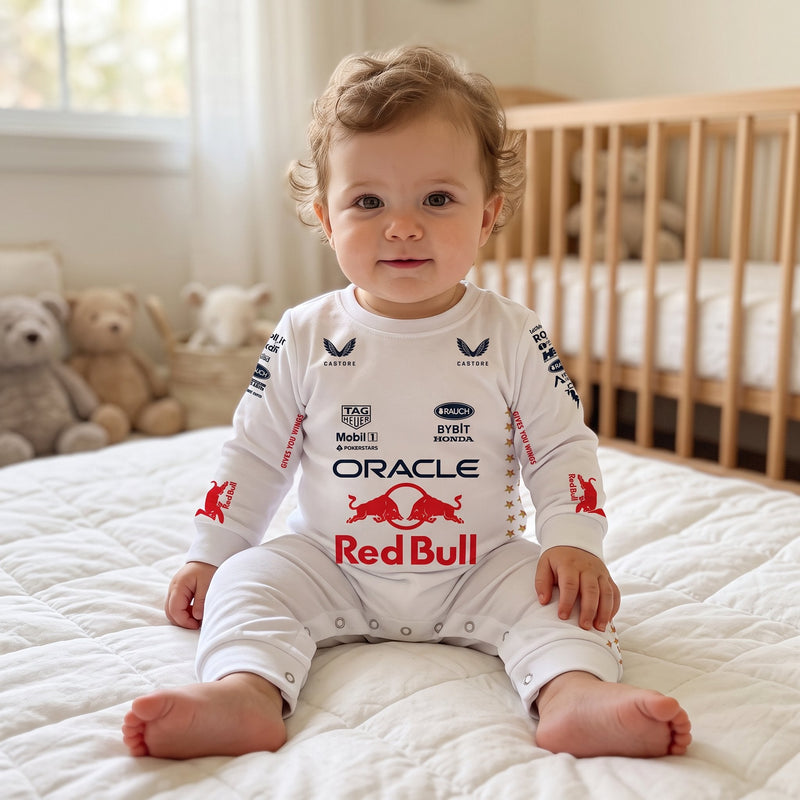 Smiling baby in a white Oracle Red Bull F1 baby onesie with team sponsor logos, sitting on a quilted bed in a nursery
