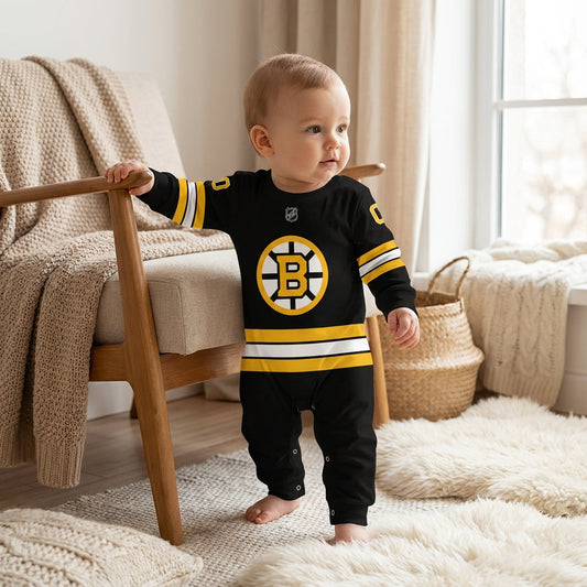 Baby wearing black Boston Bruins NHL onesie with spoked-B logo standing by nursery chair with knit blanket