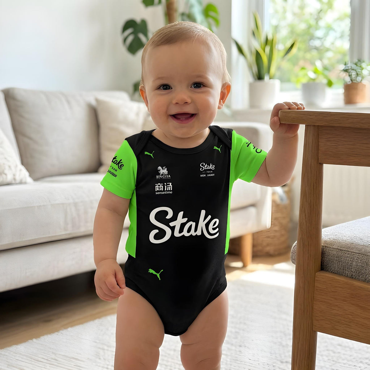 smiling baby wearing Stake F1 Kick Sauber black green bodysuit standing and holding chair in living room